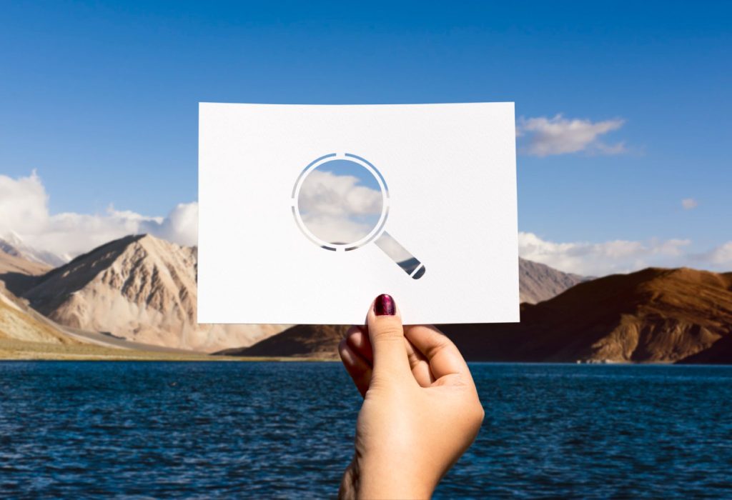 A hand holding up a cut-out magnifying glass framing a cloud above mountains.